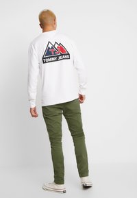 Vit långärmad t-shirt med en svart, röd och blå bergslogotyp på baksidan, kombinerad med gröna byxor och vita sneakers.