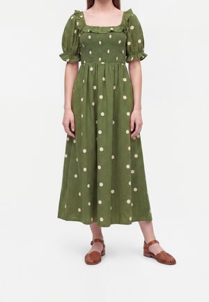 Mujer con vestido midi verde con lunares blancos, mangas cortas abullonadas, escote cuadrado con volantes y sandalias marrones de punta cerrada.