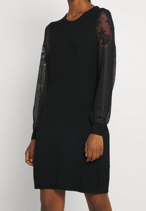 Woman wearing a black knee-length dress with sheer long sleeves featuring lace floral patterns and ribbed cuffs.