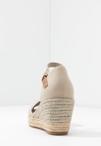 Sandalia de cuña beige con suela de cuerda trenzada, talón cerrado y tira en el tobillo con un hebilla plateada sobre fondo blanco.