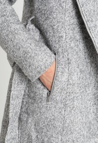 Grå texturerad kappa med hel dragkedja, försedda med en sidficka och en skräddarsydd passform. Tyget verkar tjockt och varmt.