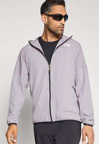 Ανοιχτό γκρι ανοιξιάτικο μπουφάν κατασκευασμένο από ελαφρύ ύφασμα, με φερμουάρ μπροστά, ελαστικά μανίκια και λογότυπο στο στήθος.