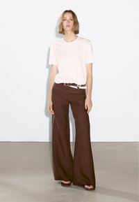 Light pink cotton t-shirt paired with brown wide-leg pants and a brown belt. Model is wearing black sandals, against a plain backdrop.