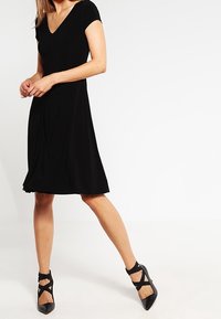 Robe noire midi avec un corsage ajusté, des manches courtes et un col en V. Associée à des talons noirs à lani ères avec des bouts pointus.