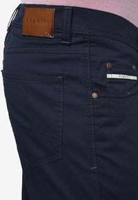 Námořnické modré kalhoty s viditelným šitím, kožená nášivka značky na pase, malá kapsa s logem, nošeny s světle růžovou košilí.