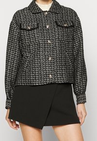 Svart och vit rutig jacka med krage, två framfickor med svart kant och silverfärgade tryckknappar. Texturerad tyg.