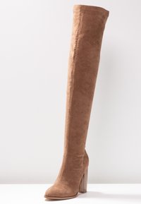 Bota de ante marrón por encima de la rodilla con un tacón en bloque, que presenta una textura suave y un diseño de punta redondeada, mostrando una silueta alta y ajustada.
