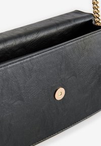 Juoda dirbtinės odos rankinė su tekstūruota paviršiaus struktūra, auksinės grandinėlės dirželis ir auksinis spaudžiamas mygtukas. Matomas vidinis atidarymas.