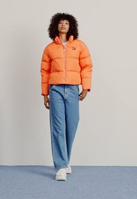 Giubbotto imbottito di brillanti arancione, caratterizzato da una chiusura con zip e un motivo a righe orizzontali, abbinato a jeans a gamba larga di un blu chiaro e scarpe bianche.