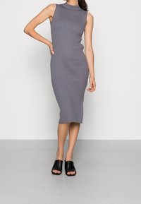 Szara dzianinowa sukienka z prążkowanym wzorem, o wysokim kołnierzu i bez rękawów, dopasowany fason, z długością do kolan. Połączona z czarnymi klapkami odkrytymi na palcach.
