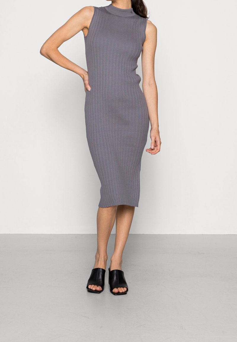 Szara dzianinowa sukienka z prążkowanym wzorem, o wysokim kołnierzu i bez rękawów, dopasowany fason, z długością do kolan. Połączona z czarnymi klapkami odkrytymi na palcach.