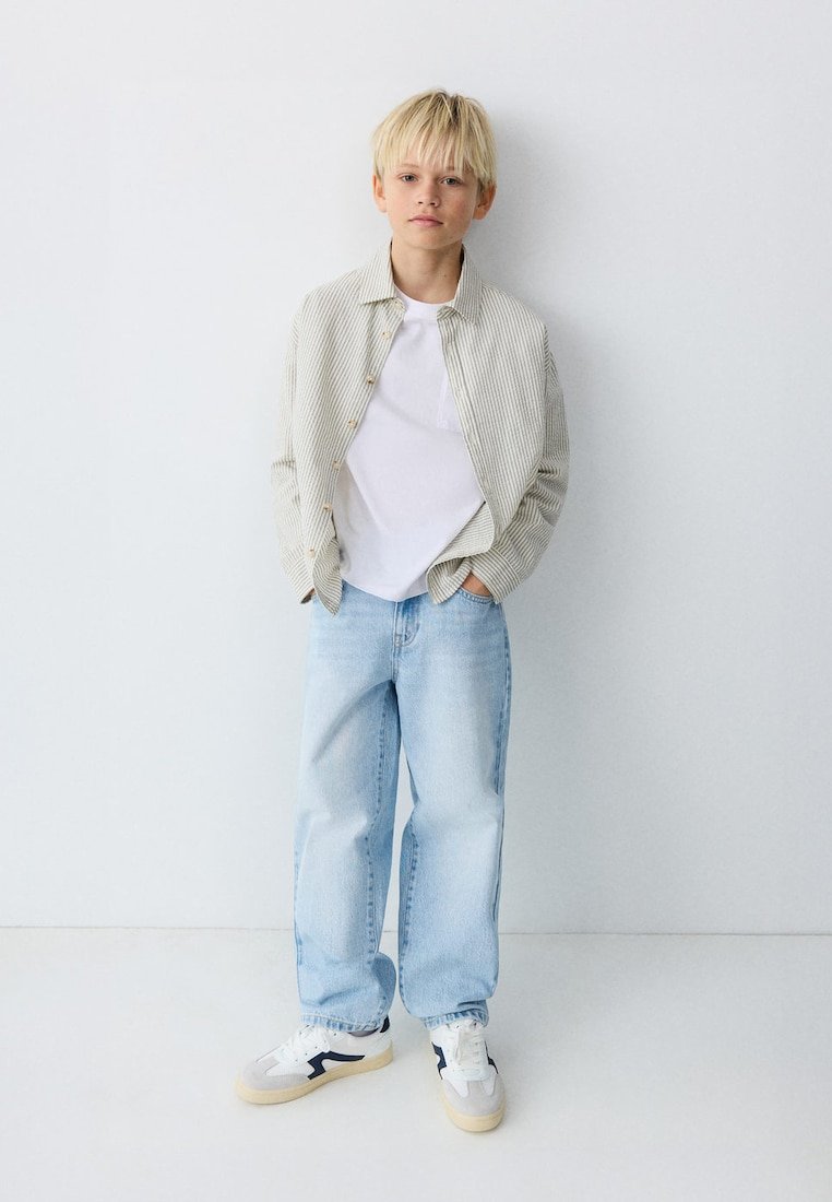 Garçon blond debout contre un mur blanc, portant une chemise beige à rayures sur un T-shirt blanc, un jean bleu clair et des baskets blanches avec des détails noirs.