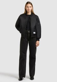 Schwarze Bomberjacke mit satinierter Oberfläche, gerippten Bündchen und Seitentaschen. Kombiniert mit hoch taillierten schwarzen Jeans und Ankle Boots.