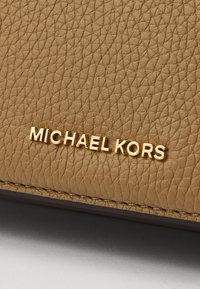 Gaiši brūnas ādas tekstūra ar akmeņveida apdari, kurai ir zelta "MICHAEL KORS" logo. Dizainā ir redzamas šuves un kompakta forma.