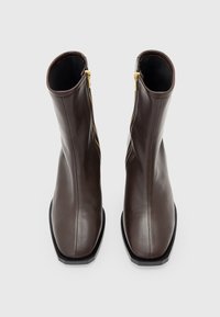 Stivali alla caviglia in pelle marrone scuro con punta quadrata e cerniere laterali in oro, esposti in posizione verticale su uno sfondo grigio chiaro uniforme.