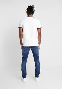 Vit pikétröja med korta ärmar och marinblå kant, kombinerad med blå jeans. Modellen visas bakifrån, med svarta dreadlocks och vita sneakers.