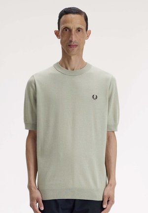 Man met kort donker haar staat recht voor met een lichtgroene korte mouw trui met een klein zwart logo op de borst.