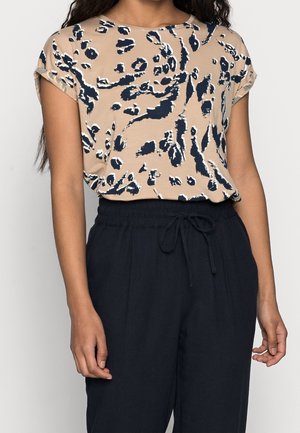 Donna che indossa un top beige a maniche corte con stampa animale in bianco e nero e pantaloni neri con cordoncino, su uno sfondo chiaro.