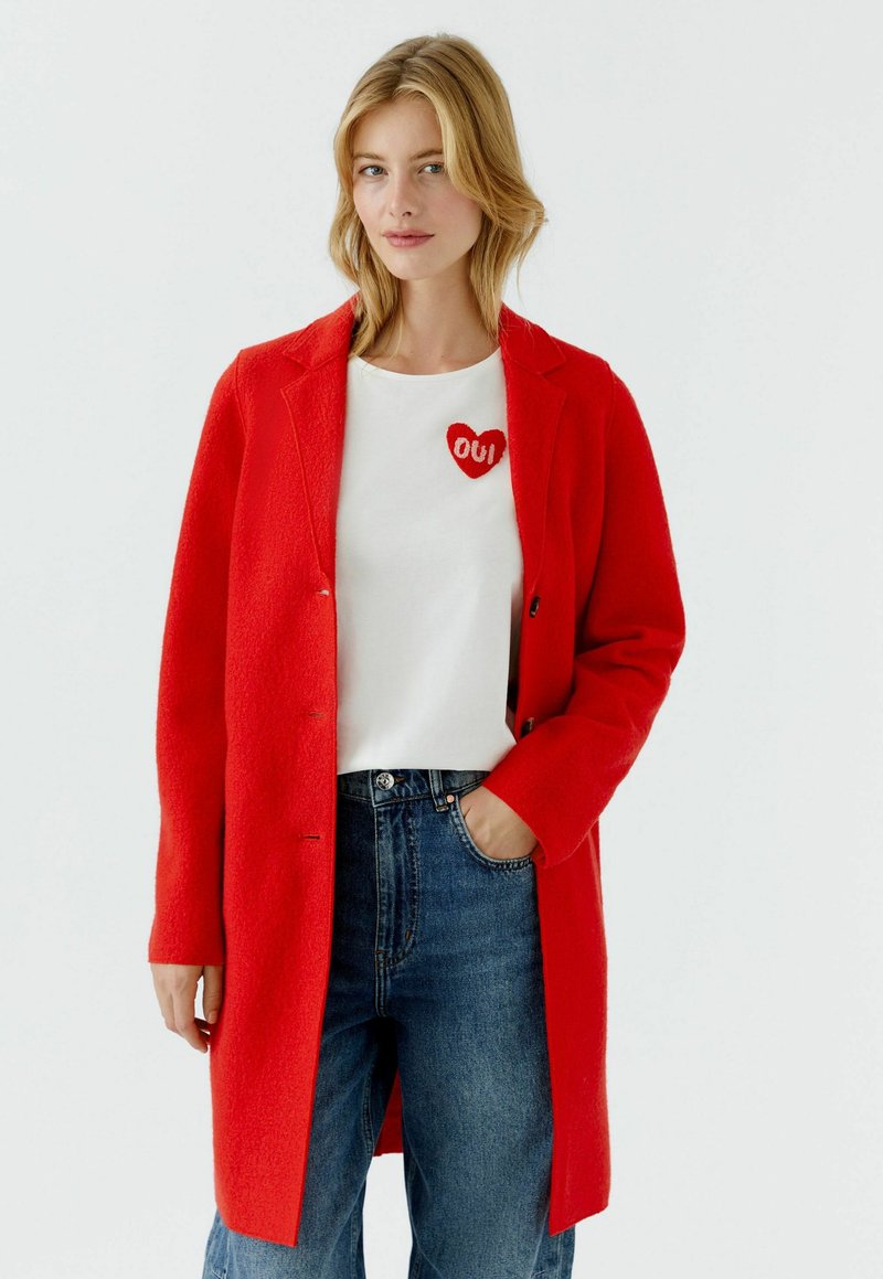 Red wool blend coat with a tailored silhouette, button front, and notched lapels. Paired with a white shirt featuring a red heart graphic.