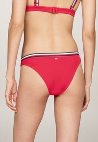 Crvena bikini donja polovica s rebrastom teksturom, koja ima kontrastnu prugastu elastiku u mornarsko plavoj i bijeloj boji. Jednostavan dizajn s minimalnim detaljima.