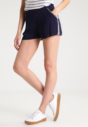 Bermudas de color azul marino con una textura suave; presentan costuras decorativas blancas a lo largo de los bolsillos y un corte relajado y holgado. Combinadas con zapatillas blancas.