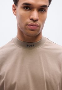 Μπεζ κοντομάνικο μπλουζάκι με στρογγυλή λαιμόκοψη και ραβδωτό γιακά. Στο μπροστινό μέρος υπάρχει μικρό μαύρο λογότυπο "BOSS". Λείο ύφασμα με μίνιμαλ σ χεδιασμό.