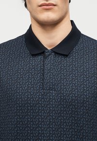 Άνδρας που φορά μπλε πόλο μπλουζάκι με ανοιχτό μπλε γεωμετρικό μοτίβο και σκούρο μονόχρωμο γιακά, εμφανιζόμενος από τον λαιμό μέχρι το στήθος.