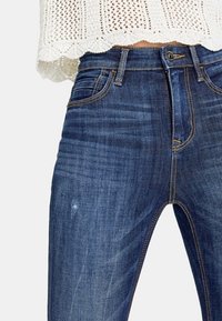 Jeans de mezclilla azul de cintura alta con un desvanecimiento ligero. Presenta un diseño estándar de cinco bolsillos y cierre de botón de metal en la cintura.
