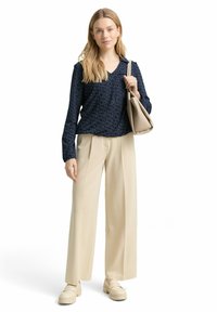 Marineblaue Bluse mit langen Ärmeln und einem dezenten Muster, kombiniert mit beigefarbenen, weit geschnittenen Hosen und hellbeigen Slip-On-Schuhen. Das Modell hält eine beigefarbene Tasche.