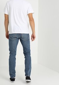 Vit kortärmad t-shirt kombinerad med blå denimjeans. Jeans har en klassisk skärning med synliga sömmar och lätt slitning. Svarta sneakers.