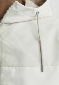Hvid zip-up jakke med ribbet tekstur, høj hals og snor med metalstoppere. Glat inderside synlig ved kraven.