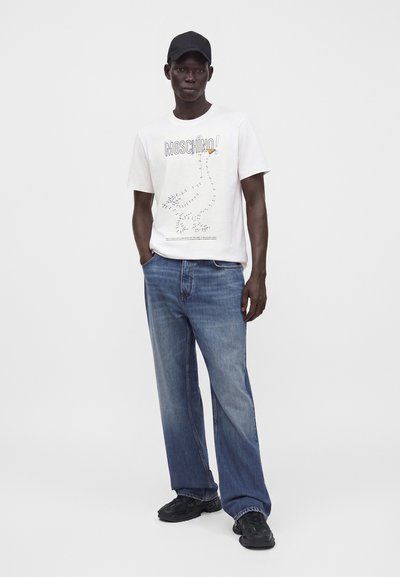 Άνδρας που φοράει μαύρο καπέλο, λευκό t-shirt Moschino με πουά σχέδιο χήνας, μπλε τζιν και μαύρα αθλητικά παπούτσια, στέκεται με το ένα χέρι στην τσέπη.