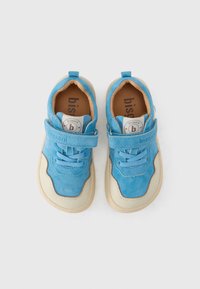 Μπλε σουέντ αθλητικά παπούτσια με κρέμ λαστιχένια σόλα· διαθέτουν λουράκι με βέλκρο, στρογγυλή μύτη και υφασμάτινες λεπτομέρειες. Στο εσωτερικό υπάρχει χαραγμένο το λογότυπο.