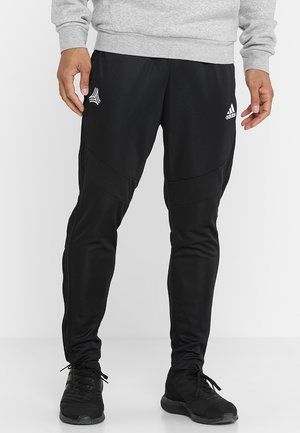 Homme portant un pantalon de survêtement Adidas noir avec des logos sur les cuisses, des baskets noires et un sweat-shirt gris, debout devant un fond uni.