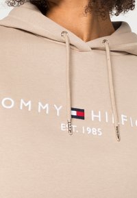 Sudadera beige con cordones, texto bordado "Tommy Hilfiger", logo de la marca y "EST. 1985", llevada por una persona con cabello rizado.