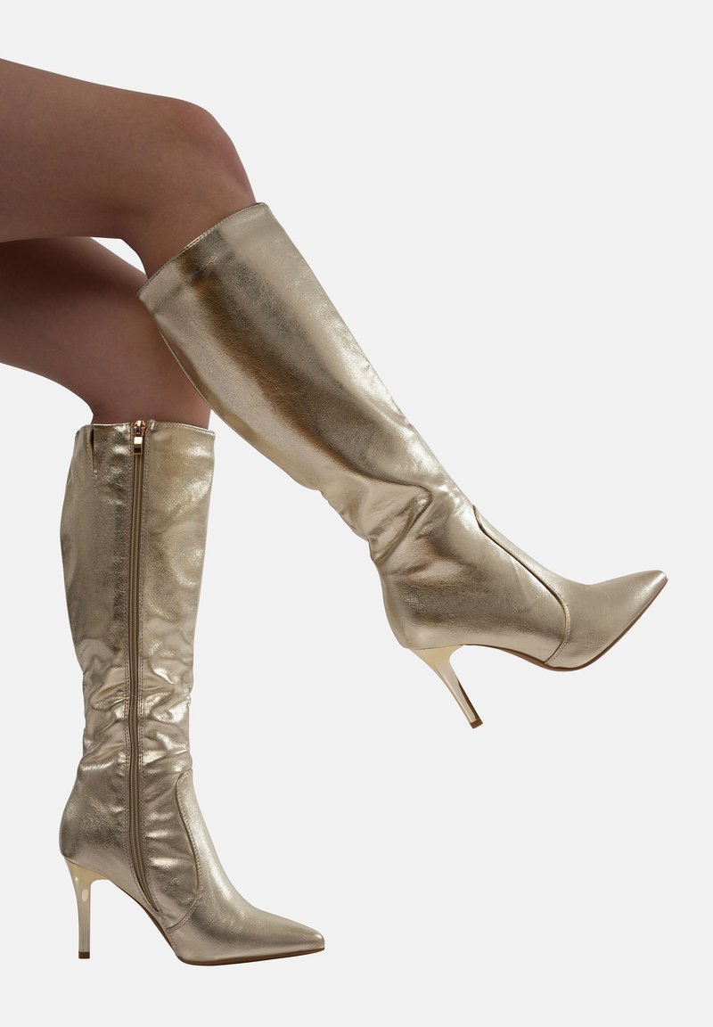 Botas metálicas doradas hasta la rodilla con punta afilada y tacón de aguja. Textura suave, cierre con cremallera en la parte posterior y diseño holgado.