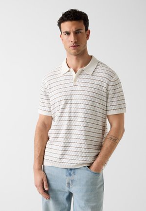 Homme portant une chemise blanche à manches courtes avec un col et de fines rayures horizontales, et un jean bleu clair, debout devant un fond blanc uni.