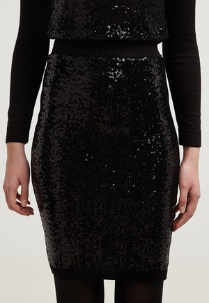 Black sequined pencil skirt worn with black long-sleeve top, hands relaxed at sides, plain light background.
