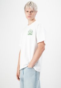 Witte katoenen t-shirt met een groene schildpadafbeelding op de borst, losse pasvorm en korte mouwen. Gecombineerd met lichtblauwe denim jeans.