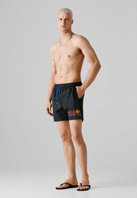 Νεαρός άνδρας με κοντά ξανθά μαλλιά στέκεται γυμνόστηθος φορώντας μαύρο σορτς Red Bull Racing και μαύρα σαγιονάρες μπροστά σε απλό γκρι φόντο.