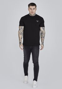 Svart bomulls-T-shirt med rund halsringning och korta ärmar, kombinerad med mörka jeans och vita sneakers. Har en liten logotyp på bröstet.