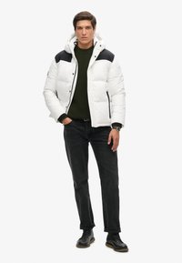 Witte pufferjack met zwarte schouders, ritsclosure en capuchon, gecombineerd met een donkergroene trui en zwarte jeans, gedragen met zwarte schoenen.