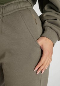 Olivgrüne Sweatpants mit weicher Textur, ausgestattet mit einer Seitentasche und einem elastischen Bund. Beinhaltet ein dezentes Markenschild.