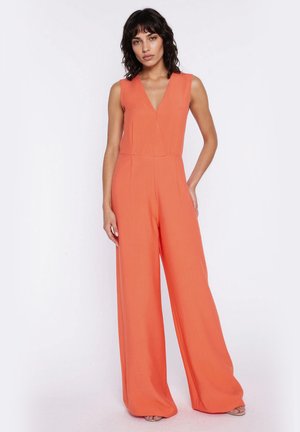 Kobieta o kręconych ciemnych włosach, ubrana w pomarańczowy, bezrękawny kombinezon z szerokimi nogawkami i dekoltem w serek, stojąca na jednolitym białym tle.