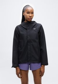 Femme aux cheveux longs portant une veste à capuche noire et un short de sport violet, debout devant un fond gris clair uni.