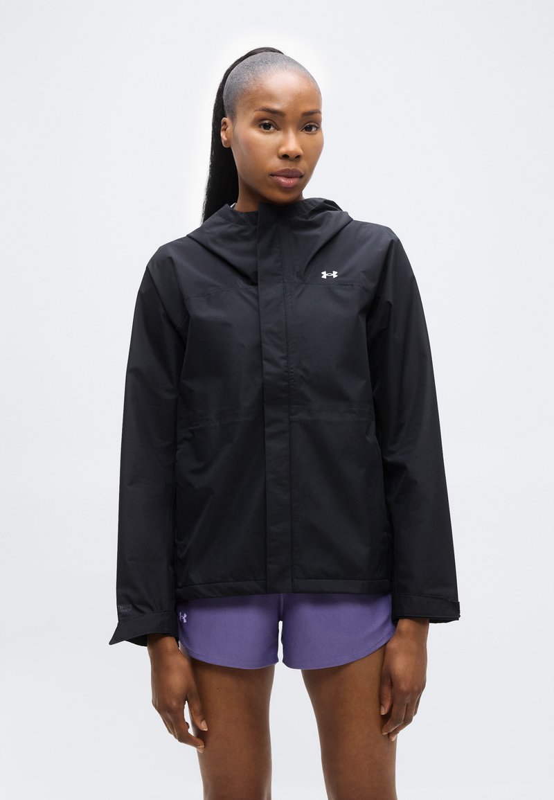 Femme aux cheveux longs portant une veste à capuche noire et un short de sport violet, debout devant un fond gris clair uni.