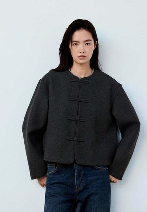 Joven mujer con chaqueta gris oscuro con botones tradicionales y vaqueros azules, de pie frente a un fondo claro y liso.