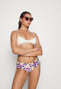 Top de biquíni branco em cruzamento, combinado com parte de baixo de biquíni com padrão floral multicolorido, apresentando laços laterais e uma textura suave. Óculos de sol transparentes.