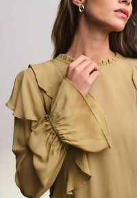 Bluză cu mânecă lungă, de culoare verde măslinie, cu accente volanate la umeri și la manșete. Material moale, ușor, cu detalii plisate la mâneci.
