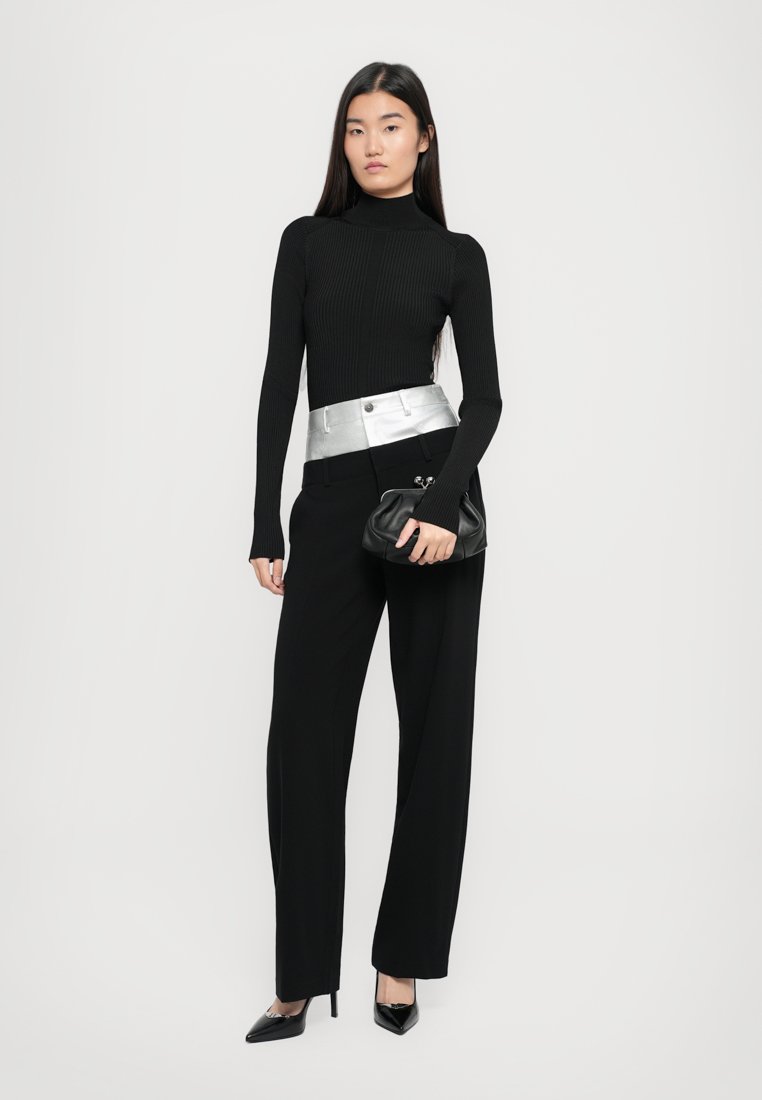Un col roulé noir à côtes, une taille ceinturée argentée, un pantalon noir à jambes larges et un sac à main noir. Le mannequin porte des talons aiguilles noirs.